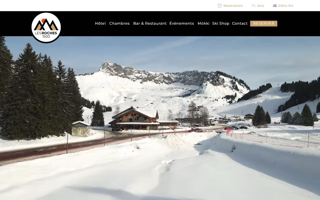 Haut de page du site de l’hôtel Les Roches 1500 : un chalet-hôtel au pied des pistes de Sommand, en Haute-Savoie, sous la neige, avec le menu et le logo de l’hôtel