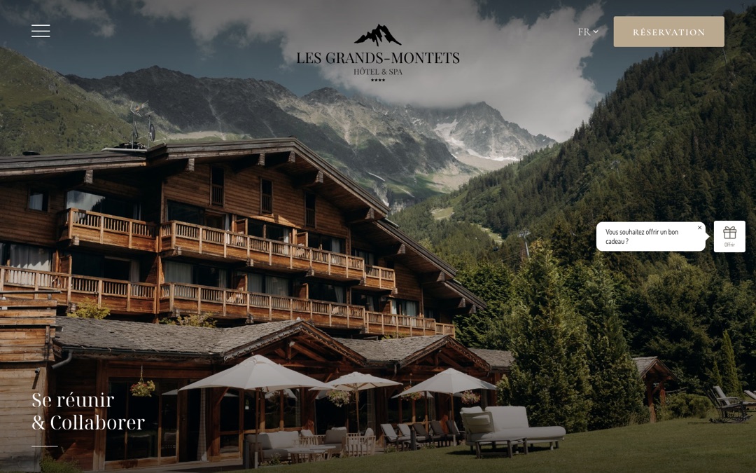 Accueil du site des Grands Montets : photo du chalet-hôtel en bois et de sa terrasse devant les montagnes, logo centré et bouton « Réservation »