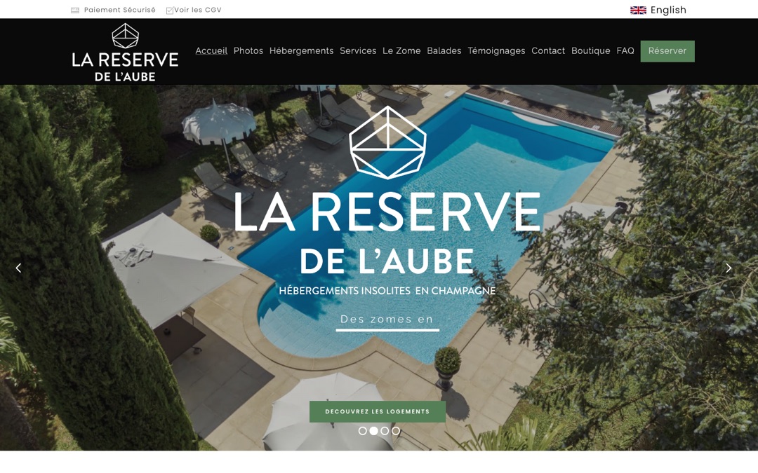 Accueil du site de La Réserve de l’Aube : vue aérienne d’une piscine entourée de parasols, logo blanc, mention « Hébergements insolites en Champagne » et bouton « Découvrez les logements »