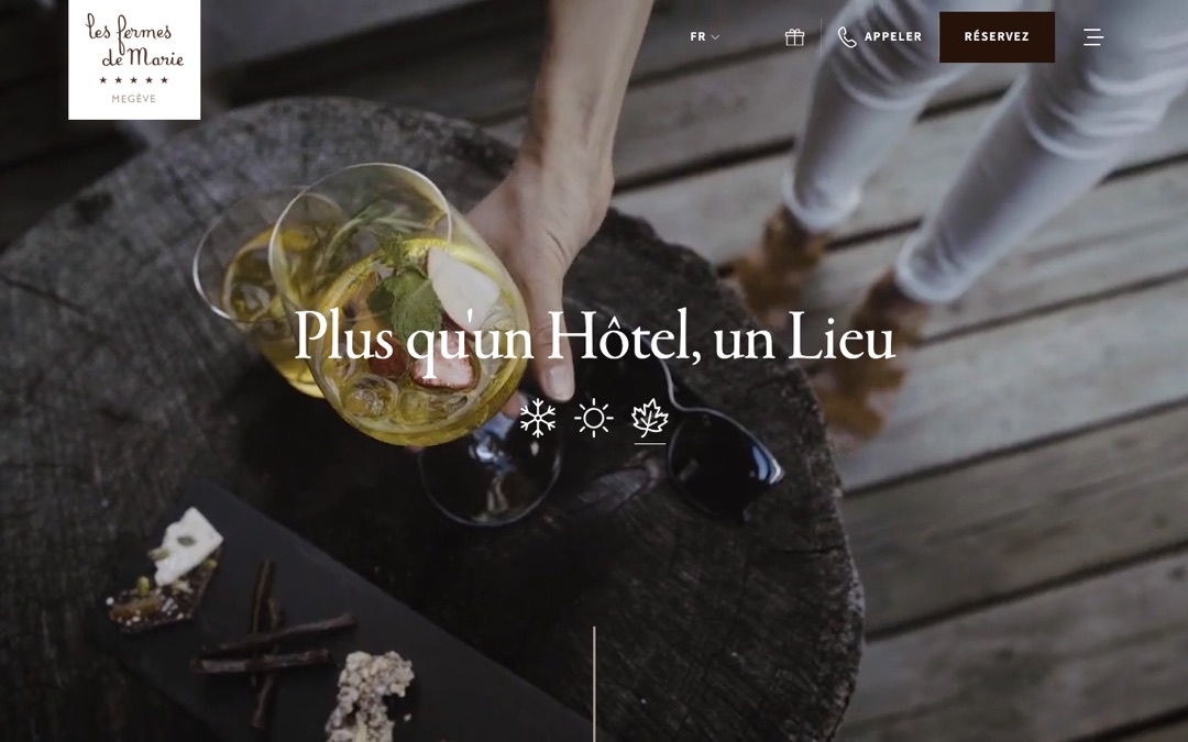 Accueil du site des Fermes de Marie : photo plein écran d’un verre et d’une main sur une table en bois, titre « Plus qu’un Hôtel, un Lieu »
