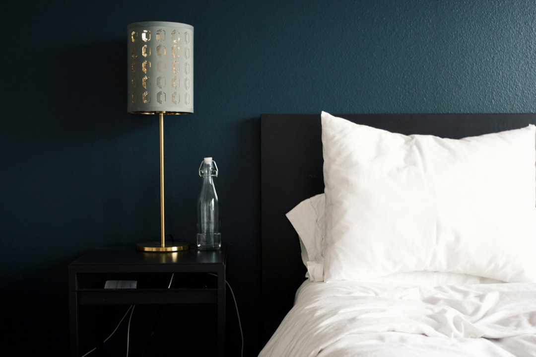 Table de chevet avec lampe et carafe à côté d’un lit aux draps blancs, dans une chambre au mur bleu nuit