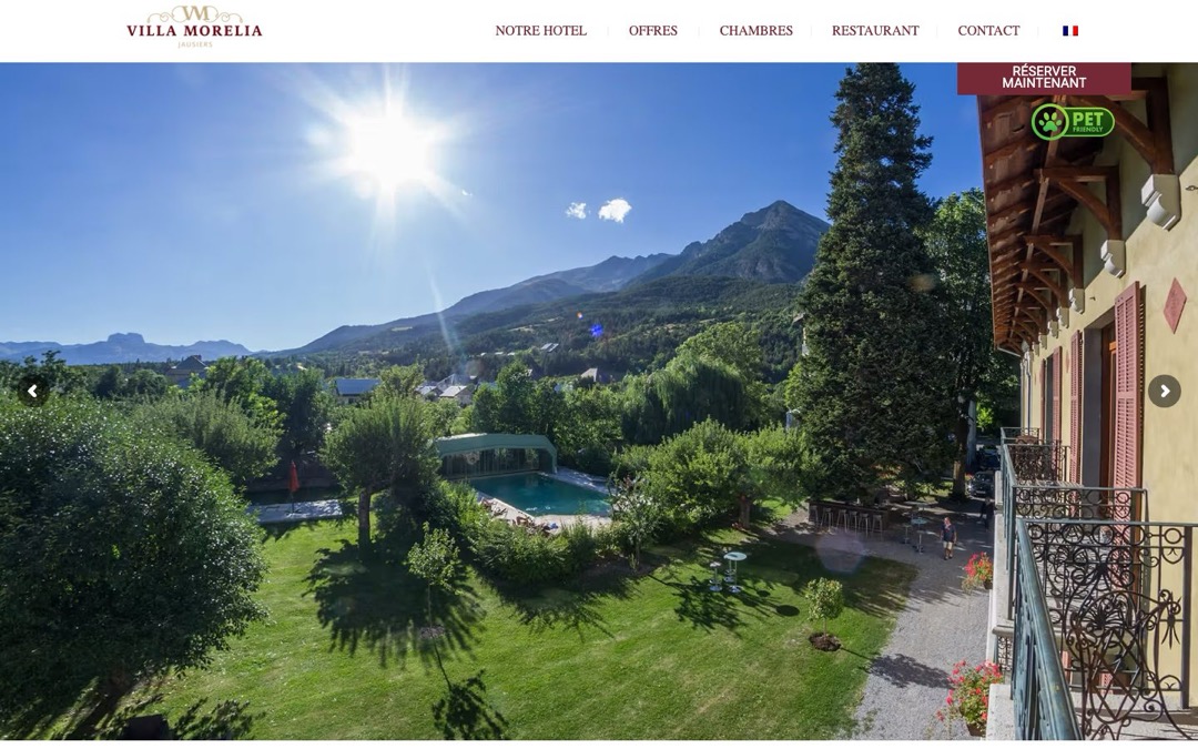 Accueil du site de la Villa Morelia à Jausiers : jardin et piscine vus d’un balcon, montagnes au fond, menu en haut de page et bouton « Réserver maintenant » en haut à droite