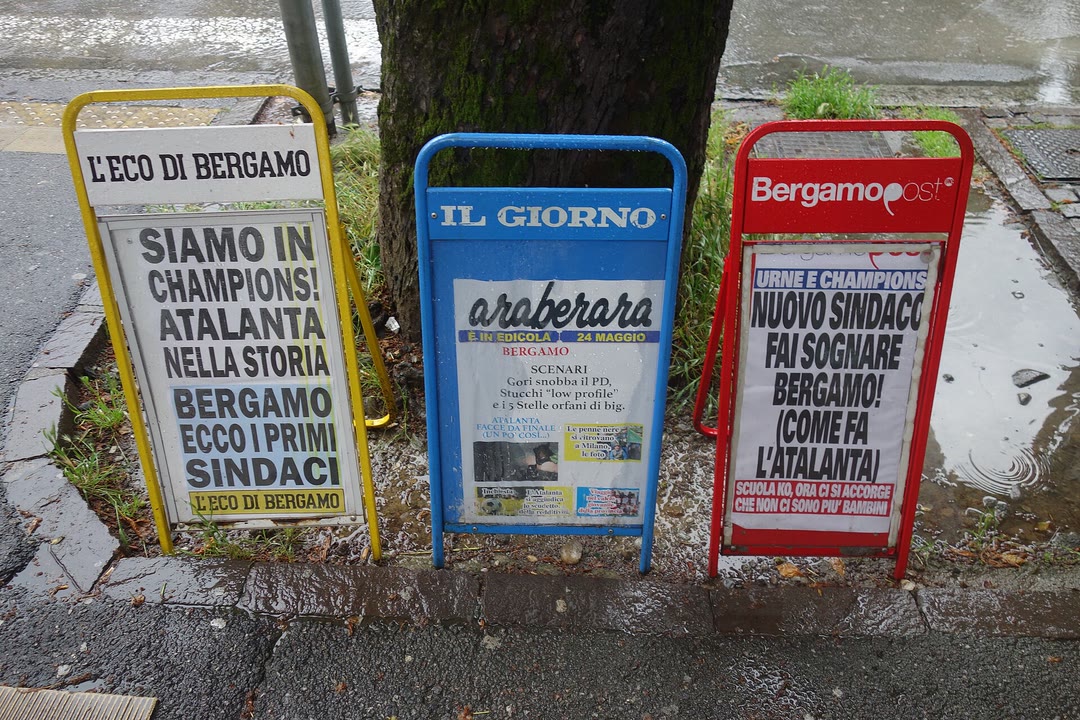 Trois chevalets de presse alignés au pied d’un arbre, sur un trottoir mouillé, affichant les unes de journaux locaux italiens