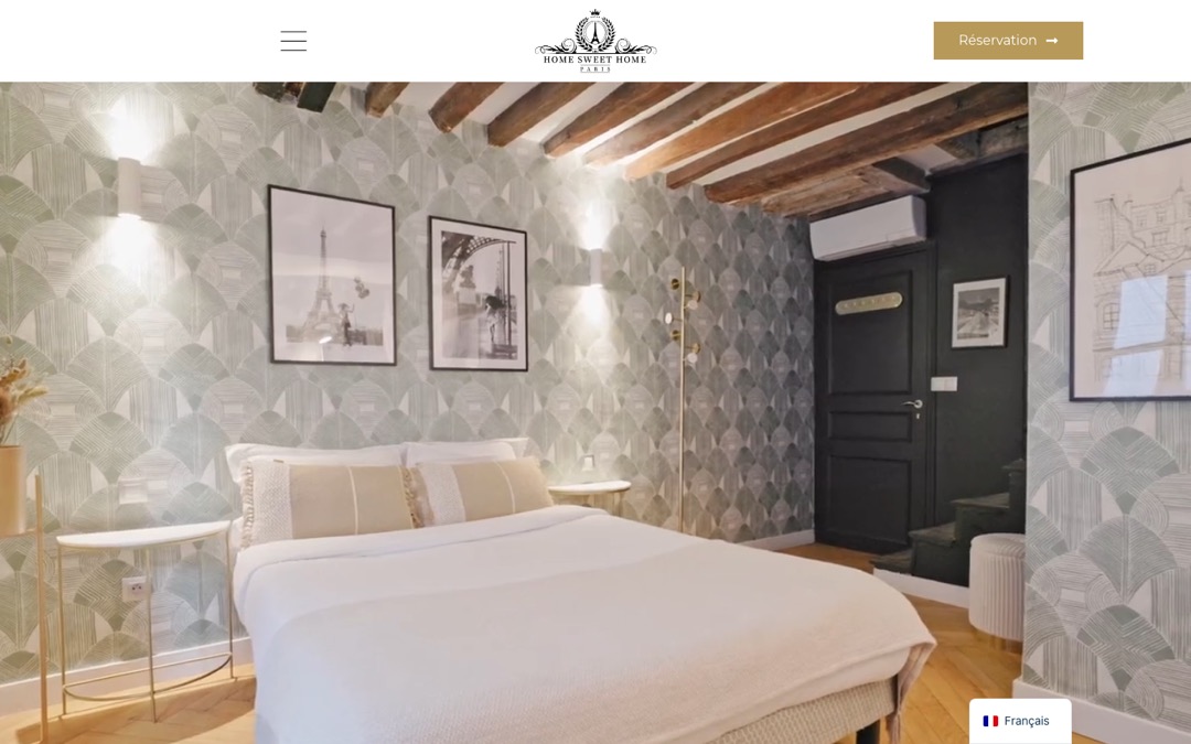 Accueil du site Home Sweet Home Paris : chambre aux poutres apparentes et papier peint vert d’eau, logo centré, menu en haut à gauche, bouton « Réservation » à droite et sélecteur de langue en bas