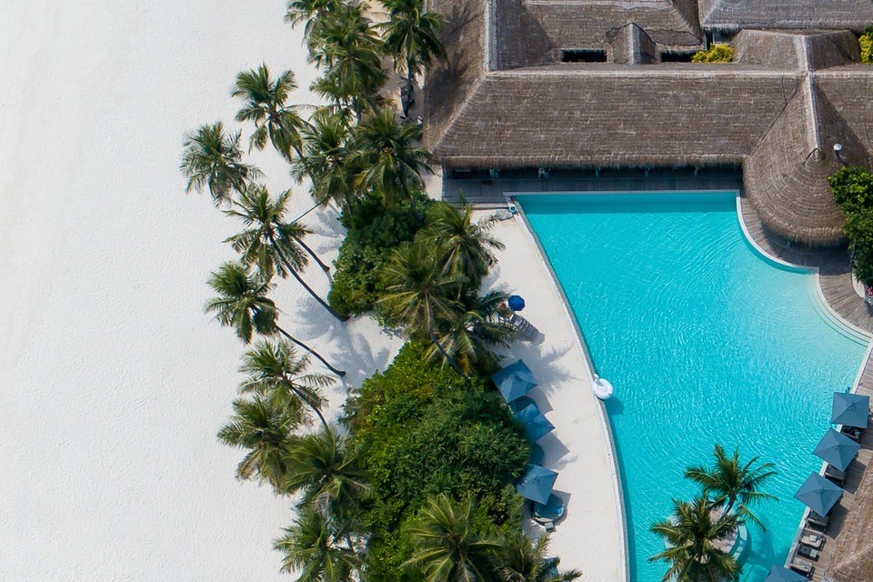 Vue aérienne : toits de chaume d’un bâtiment, grande piscine, parasols bleus, palmiers et plage de sable blanc