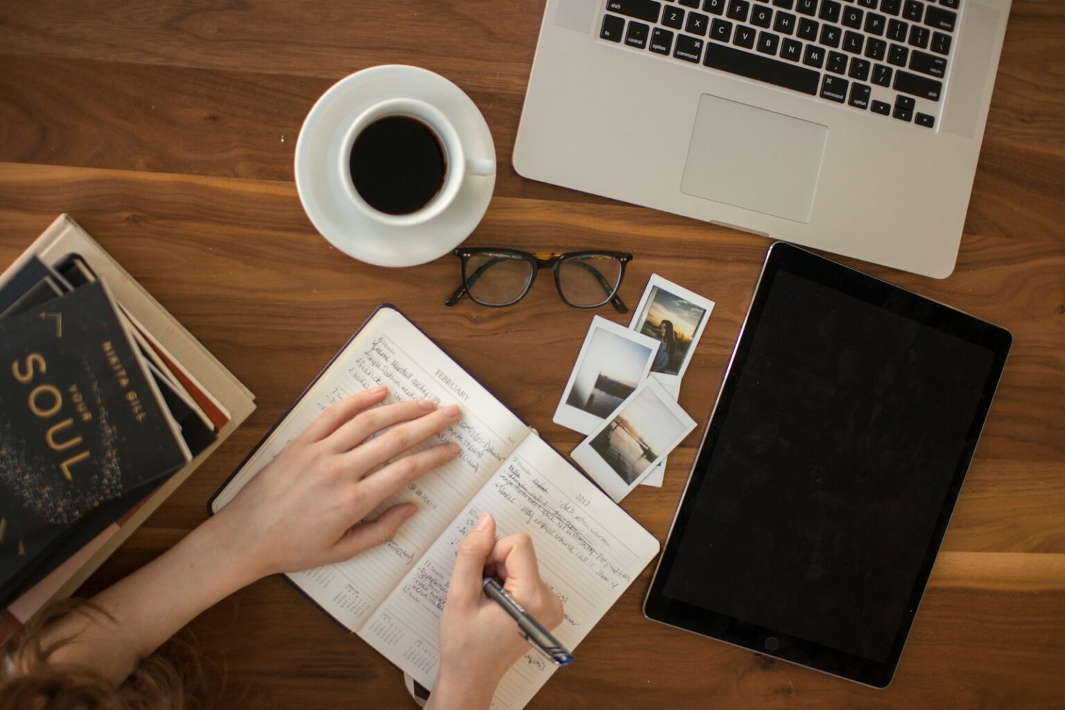 Bureau en bois vu de dessus : carnet ouvert, tasse de café, lunettes et photographies