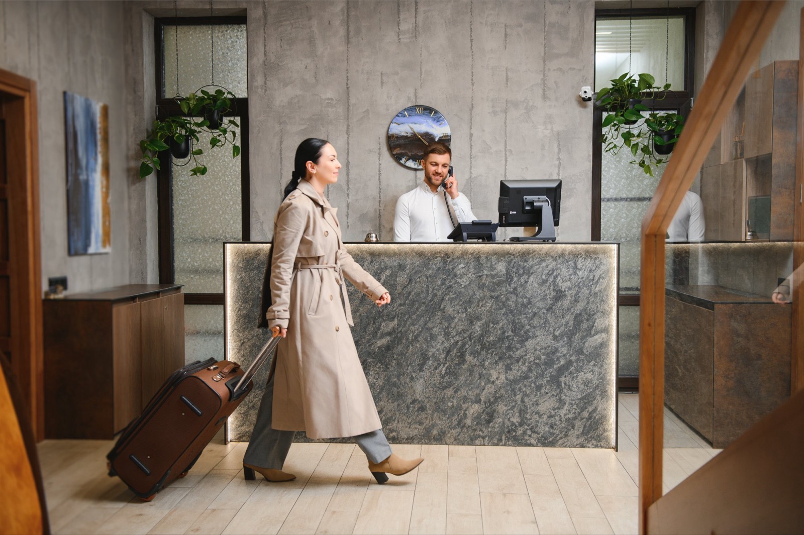 Réceptionniste d’hôtel au téléphone derrière son comptoir, une cliente passe avec sa valise