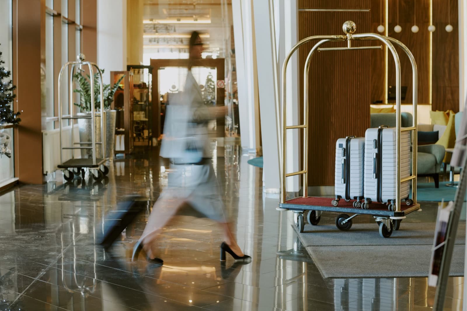 Hall d’un grand hôtel : une personne traverse le hall à grands pas, à côté d’un chariot à bagages doré chargé de deux valises