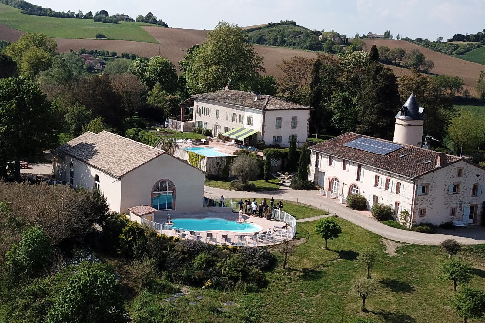Vue aérienne d’un domaine de campagne : des bâtiments anciens, une piscine et un groupe de personnes dans le jardin