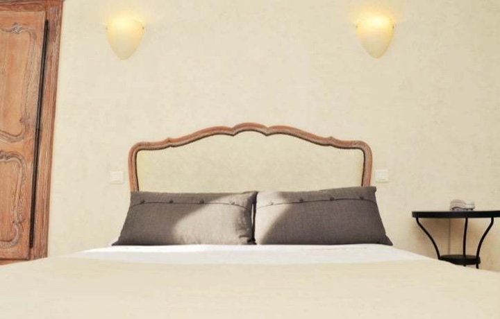 Chambre claire avec un lit à tête de lit en bois sculpté, deux coussins gris et des appliques murales