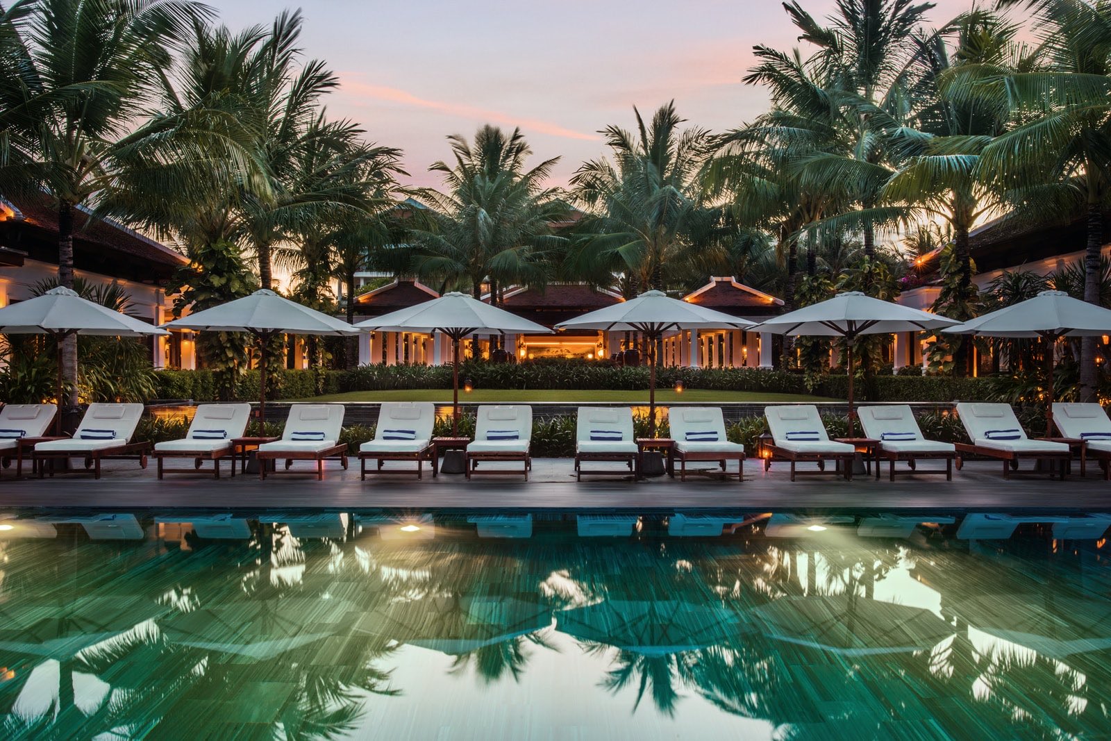 Piscine bordée de transats blancs et de parasols au crépuscule, au milieu des palmiers d’un resort