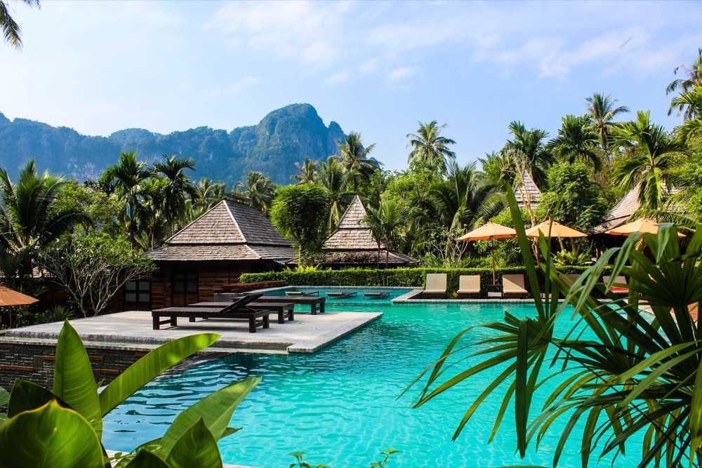 Bungalows aux toits pointus autour d’une piscine, entre palmiers et montagnes, dans un resort