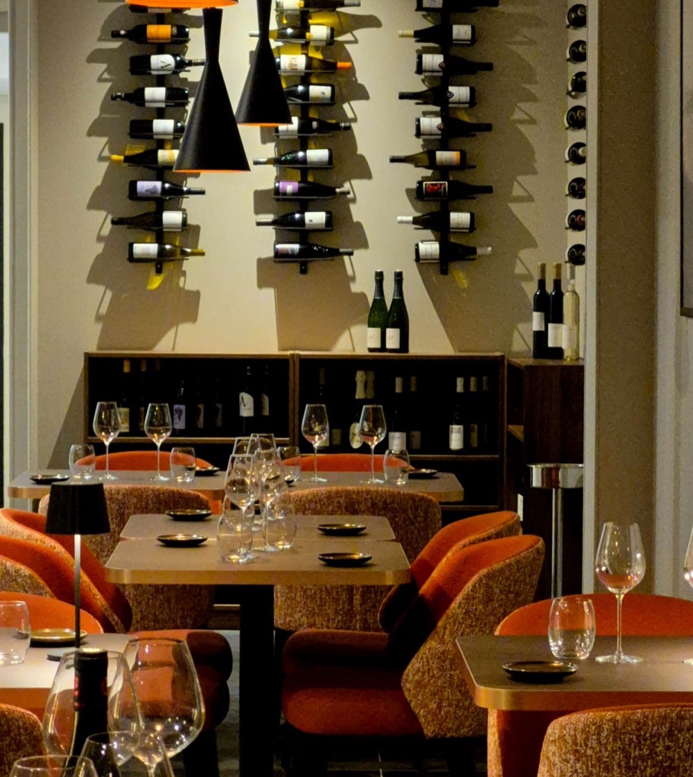 Salle d’un bar à vins : bouteilles accrochées au mur, tables en bois et fauteuils orange