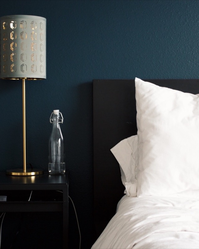 Une chambre aux murs bleu nuit, avec une lampe en laiton, une carafe d’eau et un lit aux draps blancs